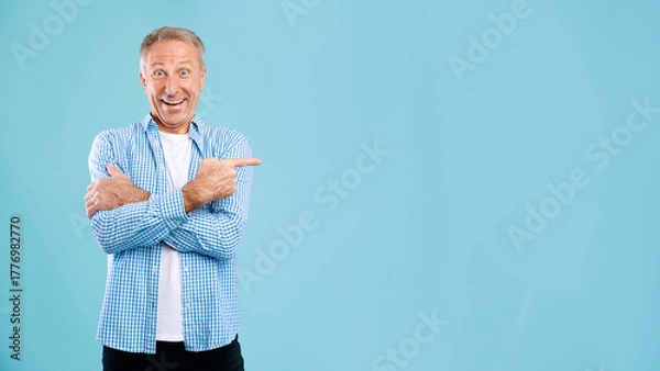Fototapeta Mature man smiles widely while pointing at the side, promoting an empty space for ads. He stands in a bright blue studio, dressed casually in a checked shirt, radiating enthusiasm.