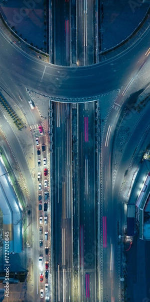 Fototapeta Aerial view of city highway interchange at night with light trails from moving cars, showing modern urban infrastructure and transportation network.