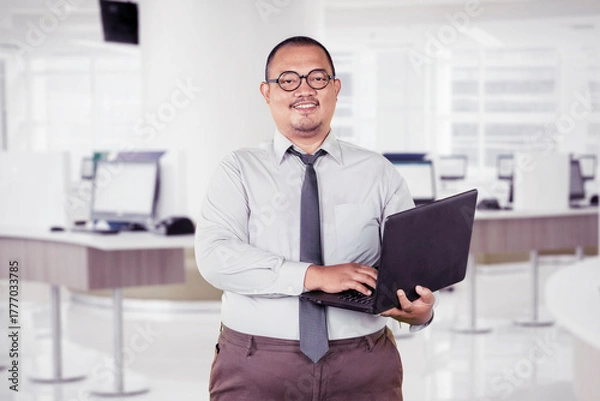 Obraz Smiling confident businessman holding and working on a laptop in a bright, modern office environment.