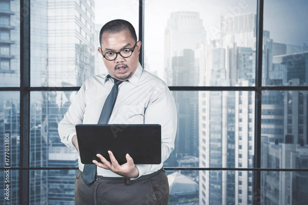 Fototapeta Shocked or Surprised Asian Businessman Staring at His Laptop in a Modern High-Rise Office