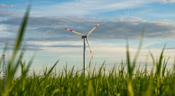 Fototapeta Wind turbine with red-tipped blades standing tall in a vibrant green field of grass under a partly cloudy blue sky in the evening light