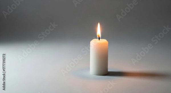 Fototapeta A white wax candle burns brightly against a neutral background, casting a soft glow and gentle shadows, perfect for representing peace, remembrance, or hope.