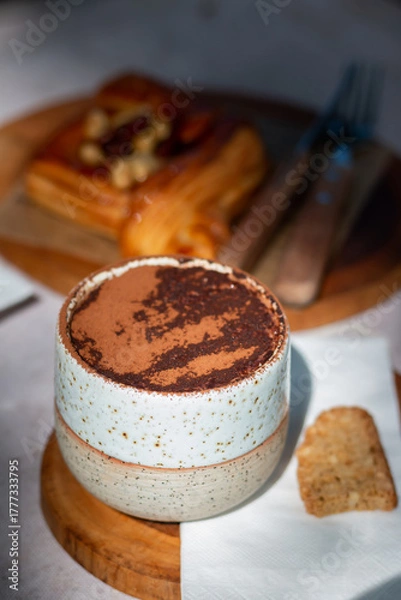 Fototapeta A cup of hot cocoa topped with cocoa powder served with pastry and cookie on wooden plate in cozy coffee shop atmosphere.