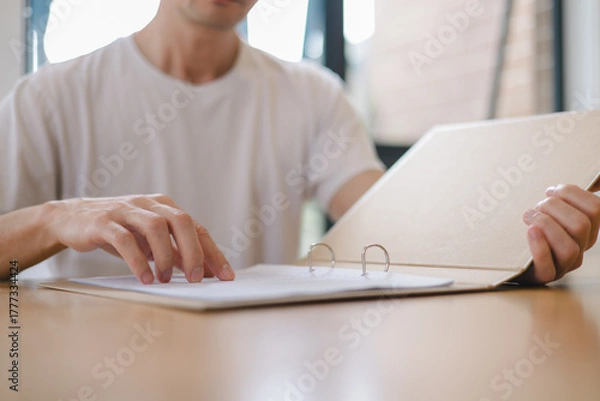 Fototapeta Close up of a man reading documents in a binder at a desk, representing office work, document review, or business analysis in a modern workspace.