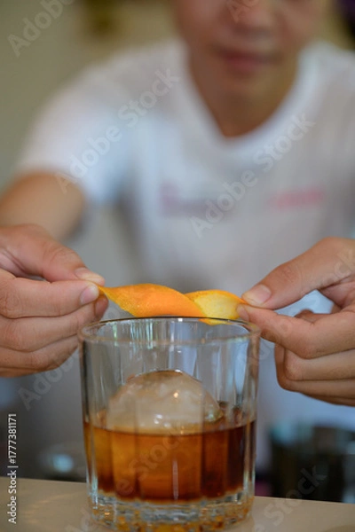 Obraz Close-up shot of bartender hands twisting orange peel over glass of cocktail with ice sphere, captured with shallow depth of field and warm soft light for premium drink concept.