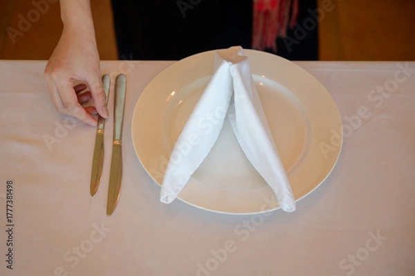 Fototapeta A close-up photo of a hand arranging silver knives beside a white plate with a folded napkin on a white tablecloth, shot in warm ambient light with shallow depth of field.
