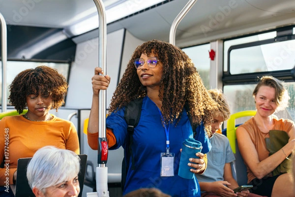 Fototapeta African female nurse wearing blue scrubs holding a water bottle and handrail while commuting on a public bus