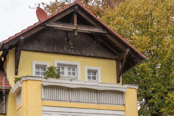 Fototapeta Charming yellow house with balcony and wooden roof details. Surrounded by green and orange autumn trees.