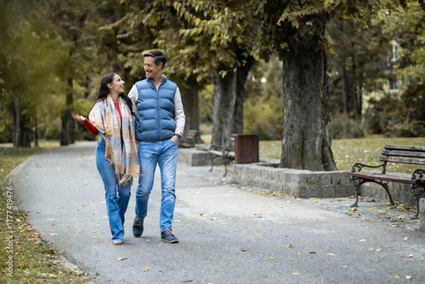Fototapeta A cheerful couple enjoys a leisurely walk in a scenic park, sharing smiles and laughter, creating a warm and inviting atmosphere surrounded by nature's beauty and colorful trees.