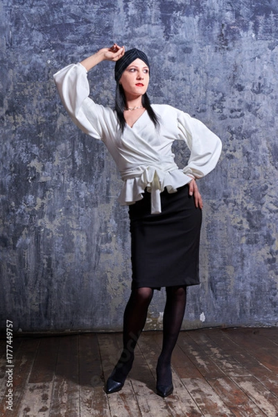 Fototapeta In a dark room stands a beautiful girl with long hair, dressed in a black leather skirt and a white blouse. Her outfit is designed in a business style. This is the image of a business woman.