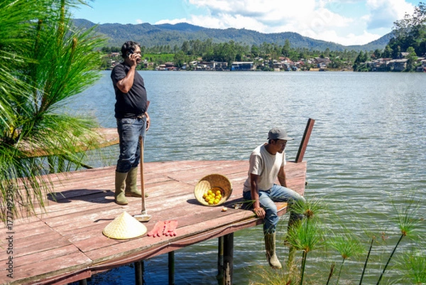 Fototapeta Two Farmers Taking Break on Wooden Dock by Lake one Chatting on His Cell Phone