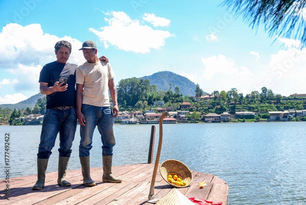 Fototapeta Two Farmers Checking Their Phone on Peaceful Lake Pier Overlooking Village on Beautiful Sunny Day