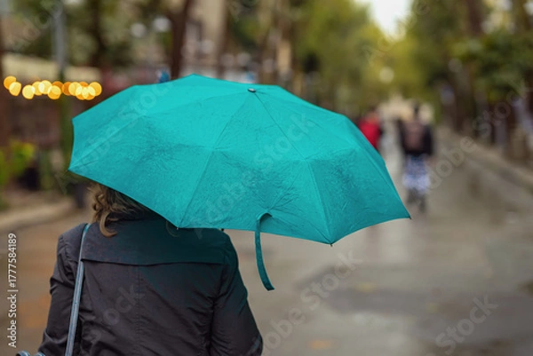 Fototapeta Woman back view with green umbrella walking on rainy city street surrounded by soft bokeh lights and blurred background