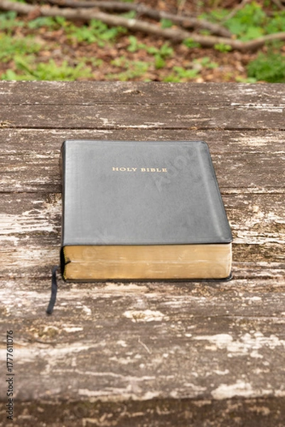 Fototapeta Holy Bible with a dark cover, gold-edged pages, and bookmark, resting alone on a textured, weathered rustic wooden table. Outdoor setting. Symbolizes faith, religion, and study.