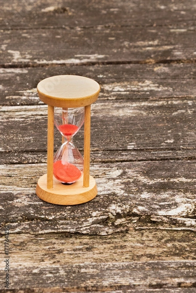Obraz Wooden hourglass with red sand running through, standing on a textured, weathered rustic wooden table. Outdoor setting. Symbolizes time, passage, countdown, and deadline.