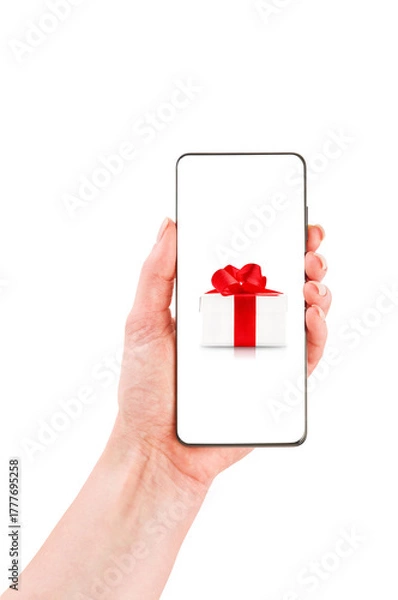 Fototapeta Female hand holds smartphone with white gift box with red ribbon on screen isolated on white background with clipping path. Design element. Winter or Christmas sales and discounts.