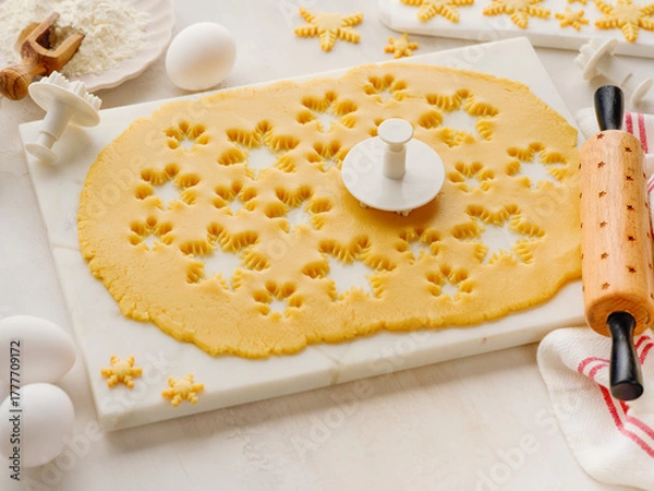 Fototapeta Rolled out dough with cut out Christmas gingerbread snowflake shapes with baking ingredients and with rolling pin on white kitchen table. Top view. Christmas baking concept background.