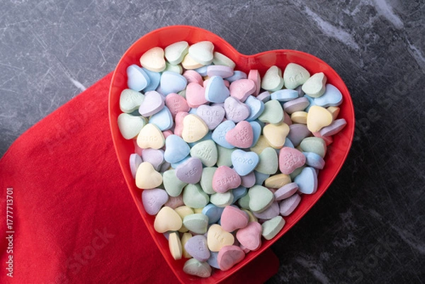 Obraz Pastel candy conversation hearts in a red heart shaped bowl on marble background with red cloth.  Valentine's Day background