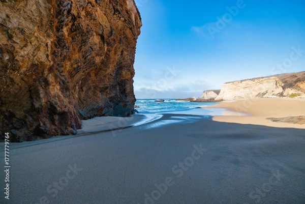 Fototapeta Scenic sandy beach with golden cliffs and turquoise ocean waves under bright blue sky showing contrast between smooth sand, textured rock formations, and gentle sea water reflections in sunlight