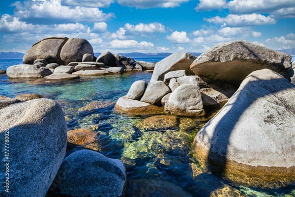 Fototapeta Rocky shoreline at Sand Harbor State Park Lake Tahoe Nevada USA with clear turquoise water sparkling in sunlight and granite boulders along pine forest coast under blue sky with white clouds
