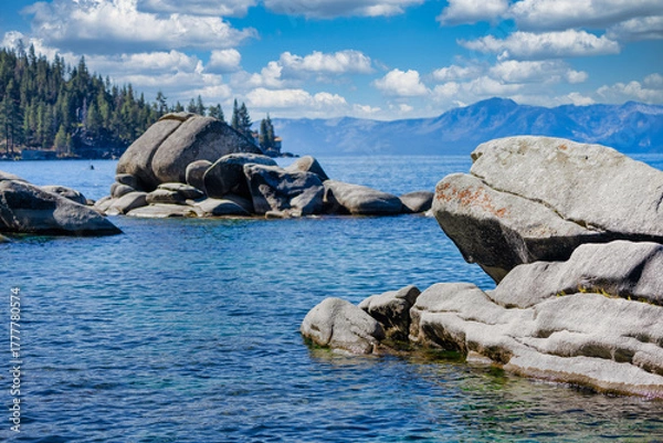 Fototapeta Granite boulders along the shore of Bonsai Rock at Lake Tahoe Nevada USA with clear blue water, pine trees and Sierra Nevada mountains under bright summer sky with white clouds on a calm sunny day