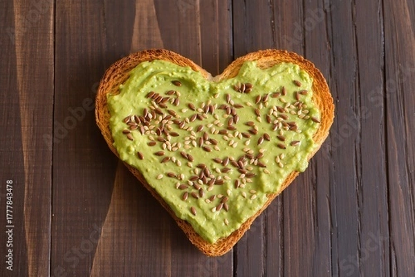Fototapeta Heart-shaped toasted bread spread with avocado paste and sprinkled with flax and sesame seeds on a dark wooden background, top view