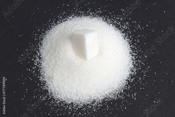 Fototapeta On a black background lies a small pile of sugar with one cube.