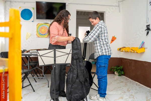 Obraz Guitar teacher helping her young student put the instrument into its case at the end of the private lesson. Music education, care for instrument, end of class, learning environment