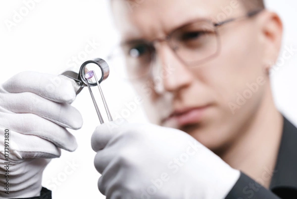 Fototapeta Male jeweler examining pink diamond with loupe and tweezers in professional setting, white studio background
