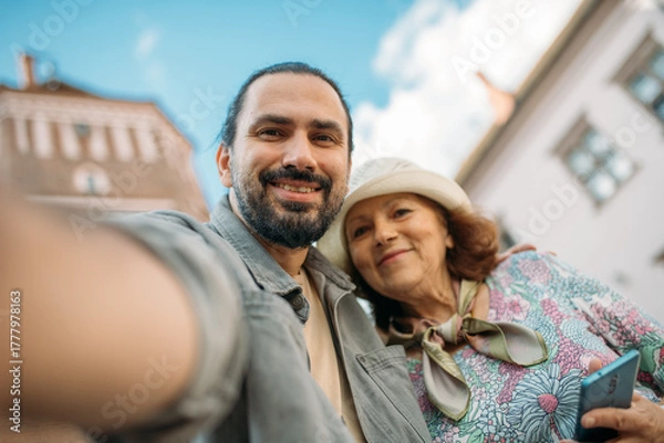 Fototapeta A pensioner and her adult son on a guided tour of an ancient castle. They smile happily and take selfies on their phones. An elderly mother and son travel together, visiting historical sites