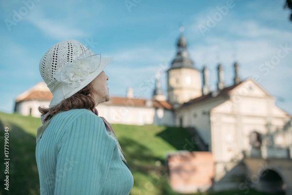 Fototapeta Пожилая довольная женщина гуляет на экскурсии в парке возле старинного замка. Пенсионерка рассматривает историческую архитектуру.