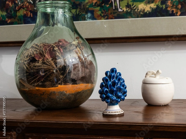 Fototapeta Close-up of decorative objects on a wooden cabinet: a glass vase with dried plants, a blue ceramic pine, and a white ceramic container. Perfect for home decor and craftsmanship themes