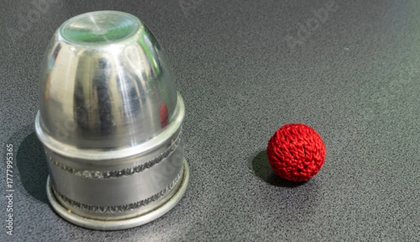 Fototapeta Metal cup and single red crochet ball on a gray surface. Minimal still life composition representing focus, illusion, and simplicity in classic magic props