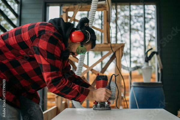 Fototapeta A young man is working with a milling cutter on the veranda of a house. The carpenter chamfers the plywood using a milling machine.