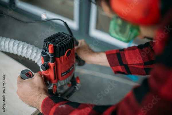 Fototapeta A young man is working with a milling cutter on the veranda of a house. Close-up of hands holding the machine. The carpenter is chamfering plywood using the milling machine.