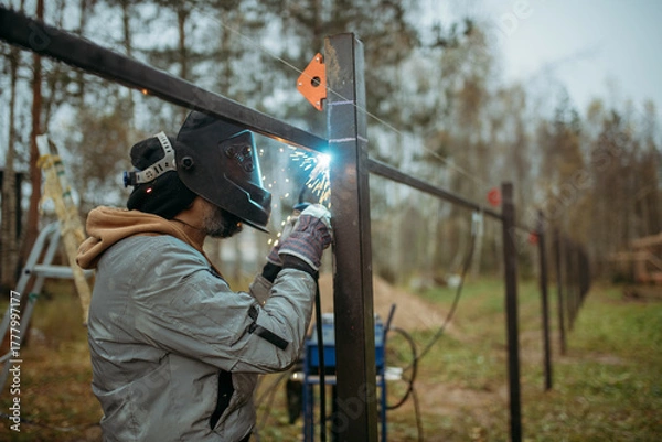 Fototapeta A young man is working as a gas welder on the street. A man wearing a protective mask is welding iron rungs to pillars. The worker, holding a welding machine, sparks fly around him.