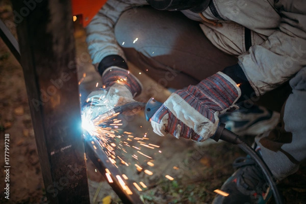 Fototapeta A young man is working as a gas welder on the street. A man wearing a protective mask is welding iron rungs to pillars. The worker, holding a welding machine, sparks fly around him.