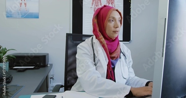 Obraz Typing Asian doctor wearing hijab, white coat, stethoscope in clinic, showing anatomical charts