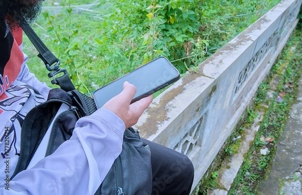 Fototapeta Person holding a smartphone while sitting on a concrete wall outdoors