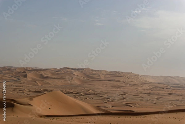 Fototapeta A vast, sweeping landscape of interlocking golden sand dunes stretching to the horizon, with a faint desert road visible in the distance