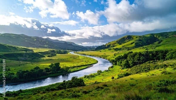 Obraz Meandering River Through Lush Green Rolling Hills Under a Dramatic Cloudy Sky with Golden Hour Sunlight