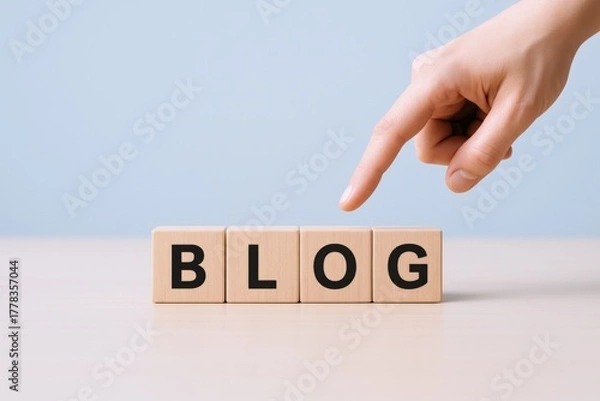 Obraz Blog Concept: Human Hand Pointing to Wooden Blocks Spelling BLOG on Table Against Blue Wall
