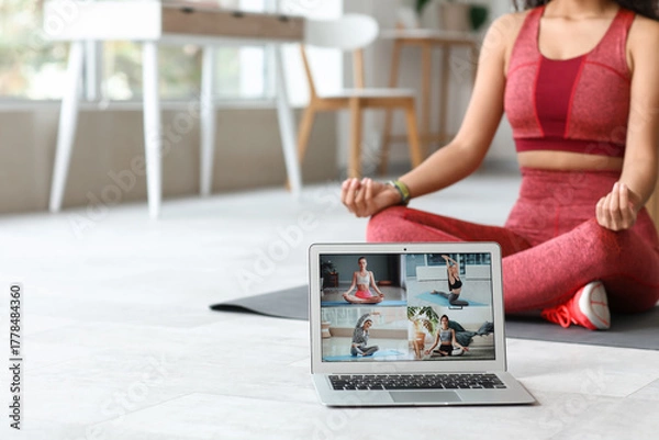 Fototapeta Sporty young African-American female coach with yoga mat and laptop meditating at home. Concept of online training