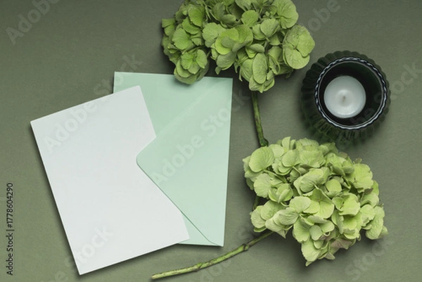 Fototapeta Blank card and envelope with two branches of greenish hydrangea and a candle on a green background. Invitation or greeting card concept.