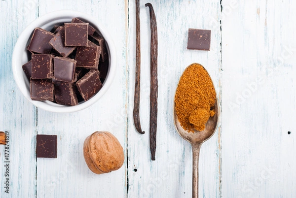 Fototapeta pile of brown sugar on old spoon, vanilla, broken dark chocolate blocks on white wooden table