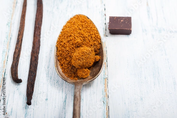 Fototapeta pile of brown sugar on old spoon, vanilla, broken dark chocolate blocks on white wooden table