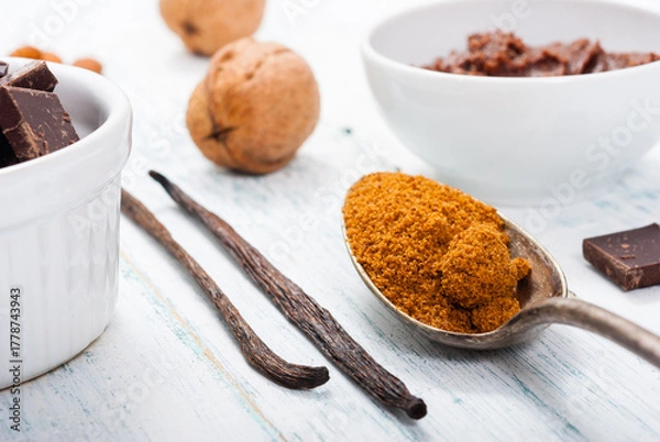 Fototapeta pile of brown sugar on old spoon, vanilla, broken dark chocolate blocks on white wooden table