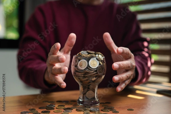 Fototapeta Close-up of hands protecting a glass filled with coins on wooden table, symbolizing financial security, savings, investment, and wealth protection concept for personal finance and money management.