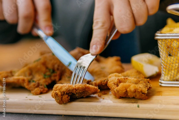 Fototapeta Close-up of a person cutting a piece of crispy, golden-brown fried schnitzel served on a wooden board with a side of french fries and lemon.