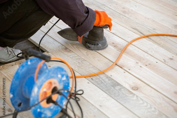 Obraz A worker in an orange glove uses an electric orbital sander to smooth a new wooden deck. An orange extension cord and a blue cable reel are on the planks.